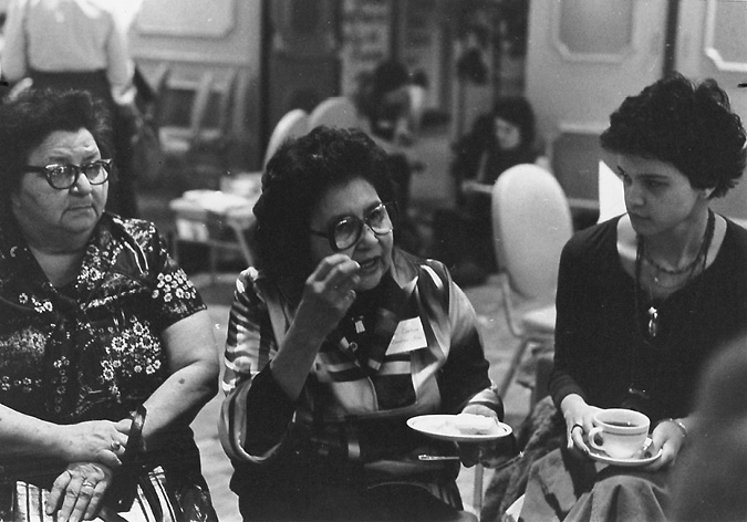 A black and white photograph of three middle aged women sitting theatre style in a room, all facing the same direction. The woman on the right is holding a cup and saucer, and the woman in the middle is holding a plate and seems to be eating.