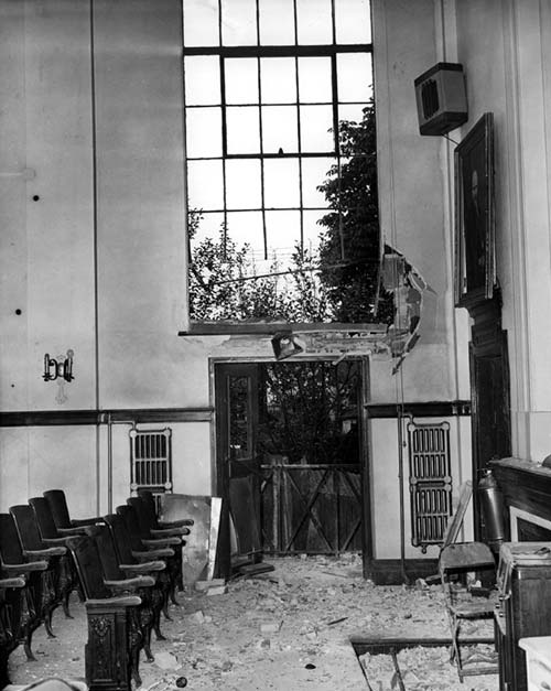 A photograph of the interior of the Labour Temple auditorium. A large window is shattered and the lower wall surrounding it is severely damaged. Theatre seating is on the left and the floor is covered with glass shards and debris from bomb blast.