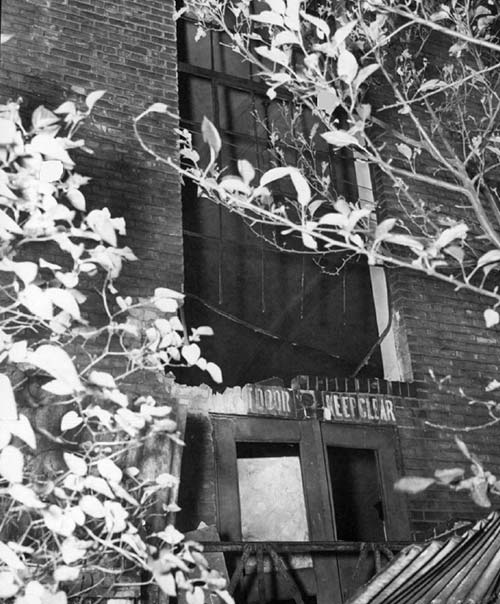 A photograph of the exterior of the Labour Temple after the bombing. There is severe damage to the window and the doors beneath. Some of the brick has been damaged and pieces are missing.