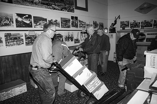 A photograph with 5 men in a room that has various large photos on the walls. One man has a dolly with 4 boxes that he is moving. There are various other boxes on the floor.