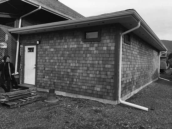A photograph of the outside portion of a building. The exterior looks to be finished with shingles and there is a pile of wood on the ground in front of the door.