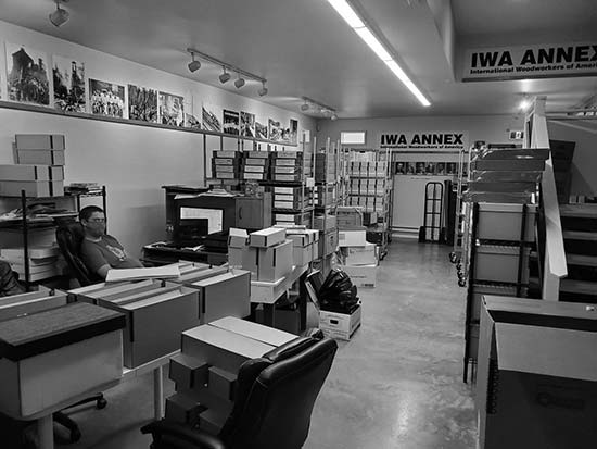 A photograph of the inside of a room. The walls have numerous photos. There are two banners up high that say, IWA ANNEX with the wording International Woodworkers of America underneath.The room is full of boxes and metal carts. There is a man sitting in an office chair.