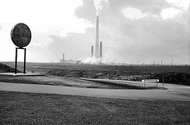 A photograph of an extremely tall smokestack, the Inco Superstack. The smokestack–one of the tallest structures of its kind–is seen in the distance encircled by its smoke and that of two smaller smokestacks which flank it.