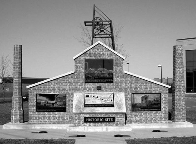 A photograph of a monument. The monument is a brick structure resembling a house. Two vertical brick columns are on either side of the building. Three panels decorate the brick: clockwise from the left, an image of a locomotive; a coal-mine scene with a pump; and an image of a coal-mine shaft. In the foreground there is an upturned panel with a map on it. Underneath the map the words historic site adorn a plaque in capital letters.