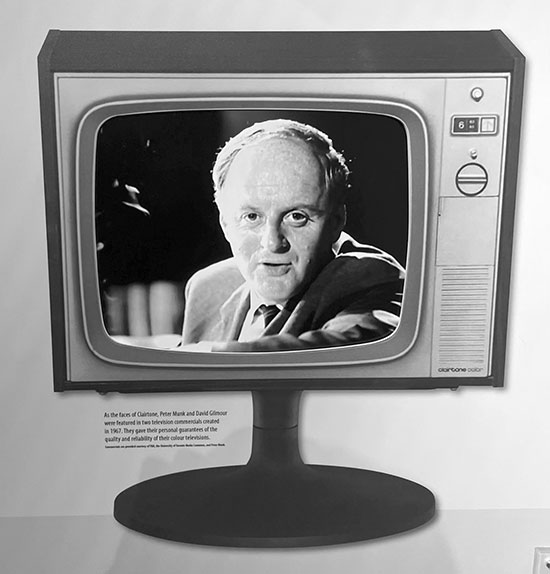 A photograph of a wall display in a museum. In a poster illustration, a man's head and face are seen staring out through a Clairtone television set that sits on a rounded base.  