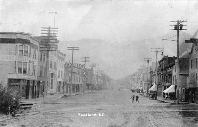 A black and white photo of street-scape in Rossland, B.C.. The road is made of dirt and there is a wagon parked on the side. The sidewalks are made of wood.