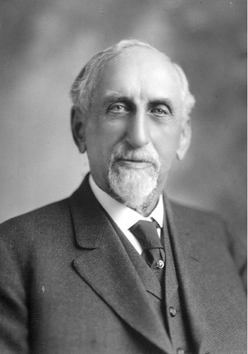 A head shot of an older white man wearing a suit. He is partially bald and has a mustache and goatee.