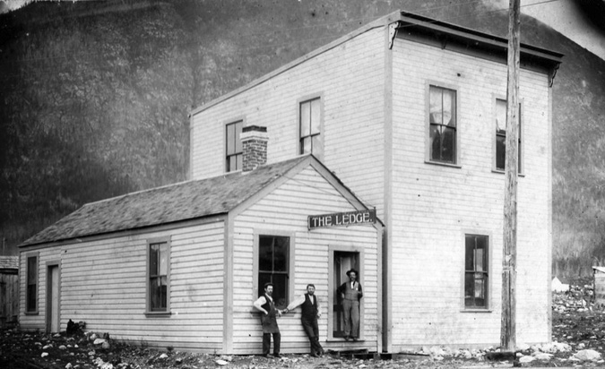 A photograph of a building with 3 men standing outside. Two are leaning beside a window and one stands int he doorway. There is a sign above the door that says "The Ledge"