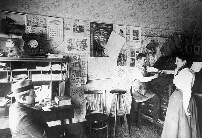 A photograph of a room with 3 people. One man with a hat sits while one man wearing an apron is standing at a printing press, and a woman is standing next to him.