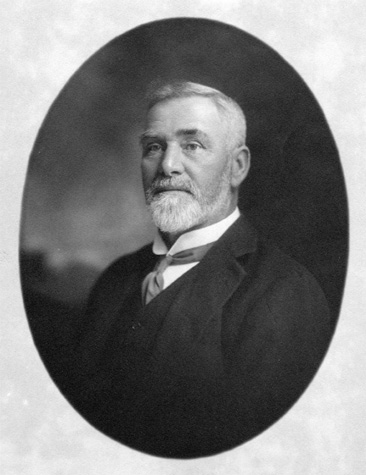 A head shot of an older white man wearing a suit. He has a gray mustache and beard.