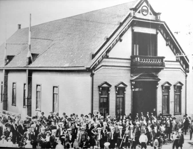 A photograph of a building with a band standing outside. The building has a balcony. The band has 75 to 100 members and they are holding their instruments.