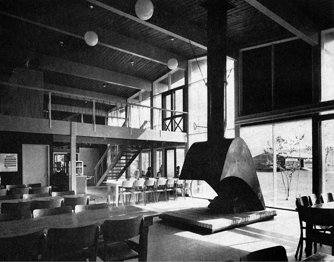 A large and airy interior, filled with tables and chairs and a large modernist fireplace, with a wall of windows looking out to the yard beyond.