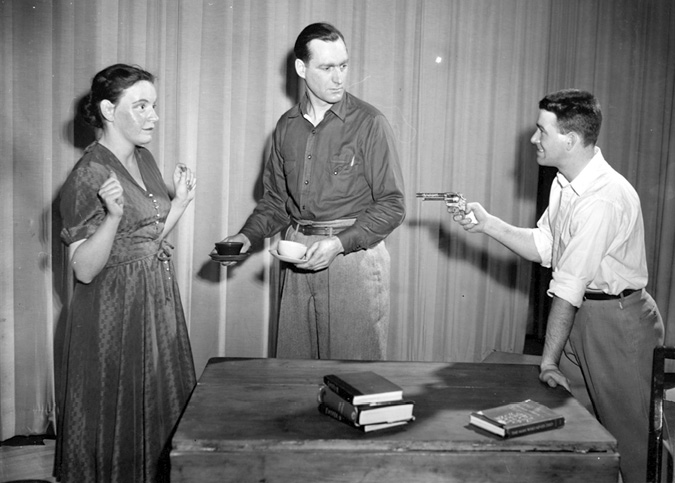 A scene from a theatre production where one man is pointing a gun, a woman has her hands up, and another man is looking on with two cups and saucers in his hands.