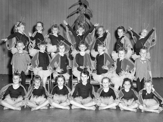 Class photo of 21 young dance students in butterfly-like costumes with antennae on their heads. The girls wear gauzy wings and tutus. The two boys are in shirts with a zigzag edge and dark pants.