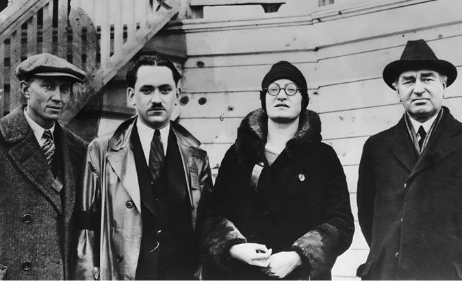 A black-and-white photo of four people standing close together, posing for the camera. They are all wearing dark, modest dresses typical of the early 20th century. There are two men on the left and one on the right with a woman standing between them.