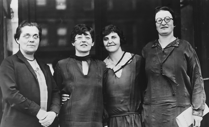 A black-and-white photo of four women standing close together, posing for the camera. They are all wearing dark, modest dresses typical of early 20th century. The two middle women are smiling, with one having her arm around the other. Their facial expressions suggest camaraderie and solidarity.