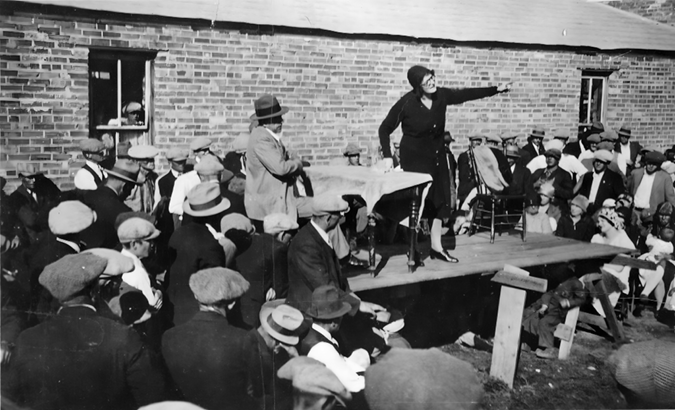 A black-and-white photo of a woman standing on a raised platform, addressing a large group of people. The woman is pointing to her left, and the crowd, mostly men wearing flat caps and fedoras, is gathered closely around her, some sitting, others standing. The setting appears to be outdoors in front of a brick building, with one person visible through a window. The atmosphere suggests a public speech or rally, with attentive listeners focused on the speaker.
