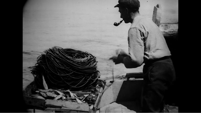 A black-and-white film-still of a man working on a fishing boat. He is wearing a flat cap and a long-sleeved shirt, with a pipe in his mouth. In front of him is a large coil of fishing rope, and fish can be seen lying on the deck beside it. The man appears to be handling the rope, preparing for or finishing a task. The background shows the open sea, and the scene captures a moment of manual labor, related to fishing.