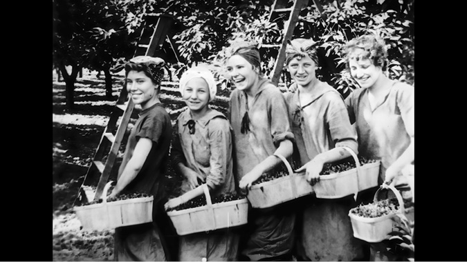 A black-and-white film-still of five young women or girls standing in an orchard, each holding a basket filled with freshly picked fruit. They are smiling and wearing headscarves, which are tied to protect their hair while working. Their clothes appear practical and slightly worn, suitable for manual labor. Behind them are trees and ladders used for reaching higher branches. The image captures a moment of camaraderie during a harvest, showcasing the hard work and community of agricultural life.