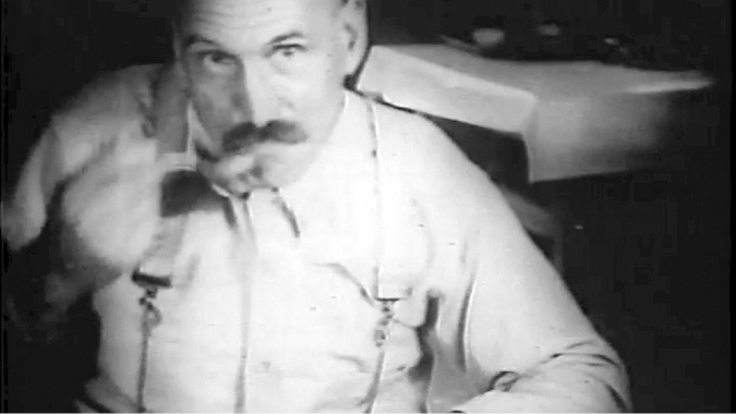 A black-and-white film-still of an older man with a mustache, wearing suspenders over a light-colored shirt. The lighting is dim, and the focus is soft, giving the image a vintage, documentary feel. The background is out of focus, but a table with a white tablecloth can be seen.