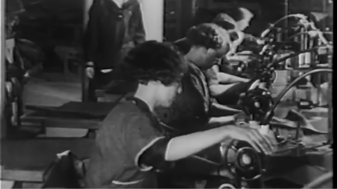A black-and-white film-still showing several women working at industrial sewing machines in a factory setting. The women are seated in a row, focused on their work. The image captures a moment from the past, with an atmosphere of labor-intensive manufacturing. You can see other workers and machinery in the background, creating a sense of a busy and productive environment. The image conveys the historical role of women in industrial labor.