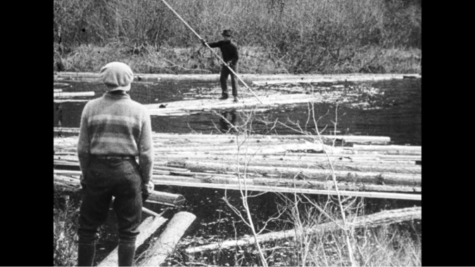 A black-and-white film-still of two men engaged in a log-driving process on a river. The man in the foreground is standing on the bank, facing away from the camera, wearing a striped sweater, trousers, and a flat cap. In the background, another man is balancing on the floating logs, using a long pole to guide them down the river. The scene captures the rugged, physically demanding work of log drivers, transporting logs across waterways.