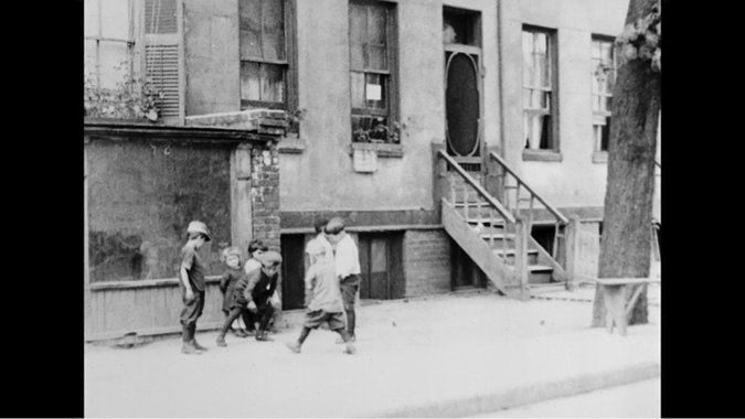 A black-and-white film-still of a group of young children playing on the sidewalk in front of a building. The children, dressed in old-fashioned clothing typical of the early 20th century, are engaged in conversation or a game. The building behind them has a staircase leading to a door with an oval window, and there are a few windows with flower boxes. The scene evokes a simple, everyday moment in an urban neighborhood, highlighting the innocence of childhood.