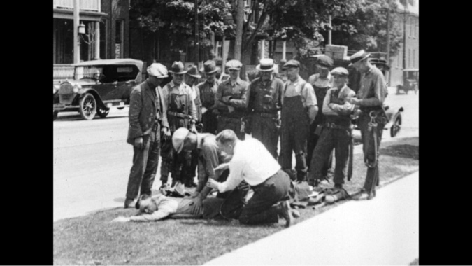 A black-and-white film-still showing a group of men, likely from the early to mid-20th century, gathered around two men tending to a person lying on the ground. The men, dressed in work clothes such as overalls and hats, appear to be observing or assisting. The person on the ground seems unconscious or injured, with two men kneeling beside them, possibly administering aid. The scene takes place on a street with vintage cars parked in the background, along with buildings and trees, suggesting it might be a public or residential area.